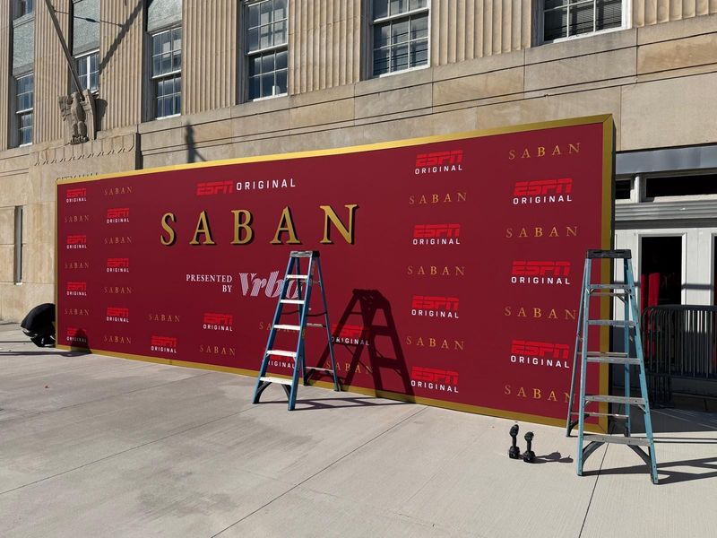 Large gold-framed step-and-repeat banner wall being installed on a sidewalk, with two ladders still standing in front of it