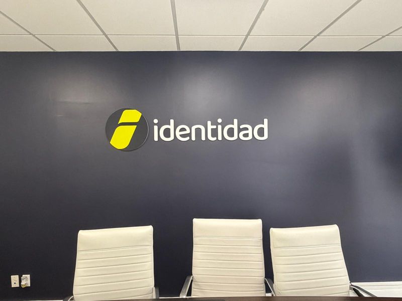 Dimensional identidad logo, a yellow and charcoal circle mark with white lettering, on a navy wall behind three white conference chairs