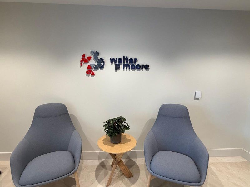 Walter P Moore dimensional logo in red, gray and navy pieces on a light gray reception wall, above two armchairs and a small side table