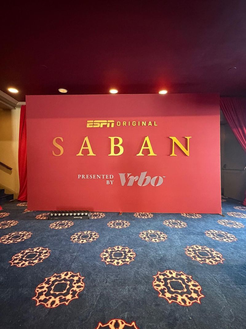 Red freestanding backdrop with raised gold ESPN Original Saban lettering and a Vrbo logo, set in a lobby lined with red drapes