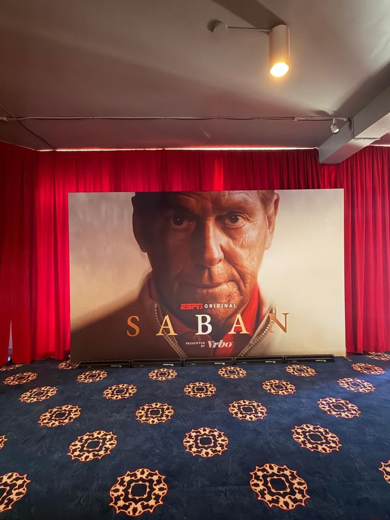 Large printed portrait backdrop for the ESPN Original Saban, standing between red velvet drapes on patterned carpet