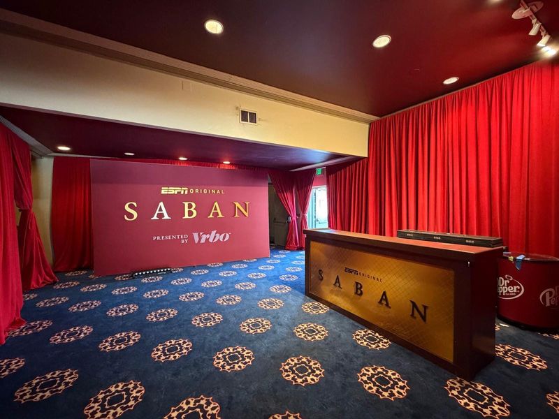 Red ESPN Original Saban backdrop with gold lettering and a matching branded bar counter in a room lined with red drapes