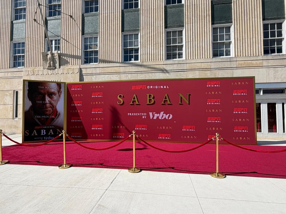 Red ESPN Original Saban step-and-repeat backdrop with a framed portrait panel, a red carpet and brass stanchions in front of a stone building