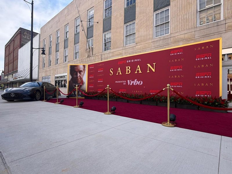 Angled view of a gold-framed Saban step-and-repeat wall with a red carpet, brass stanchions, flower boxes and carpet uplights along a sidewalk