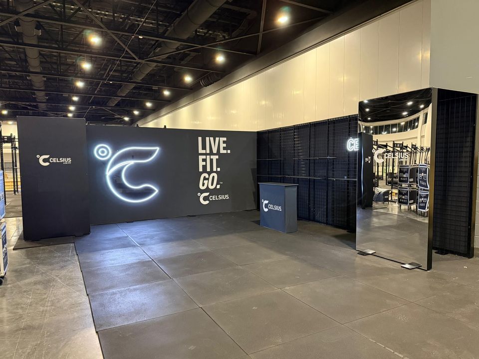 Celsius exhibit space in an exhibition hall with a dark backwall, an illuminated logo outline, Live. Fit. Go. lettering, a branded counter and black grid walls