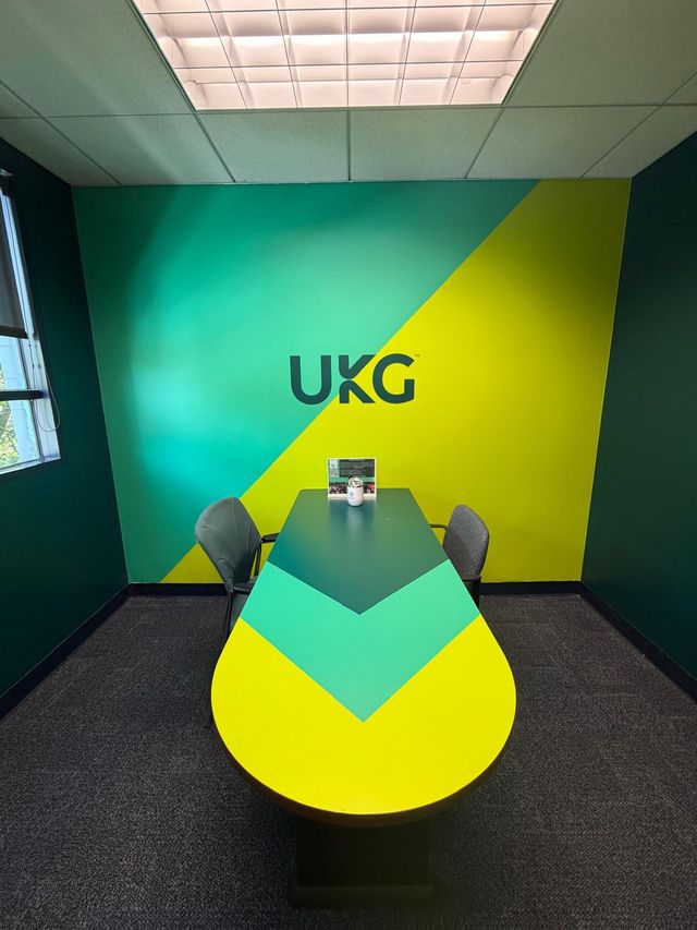 UKG-branded conference room with a teal and yellow diagonal wall mural and a matching color-blocked table